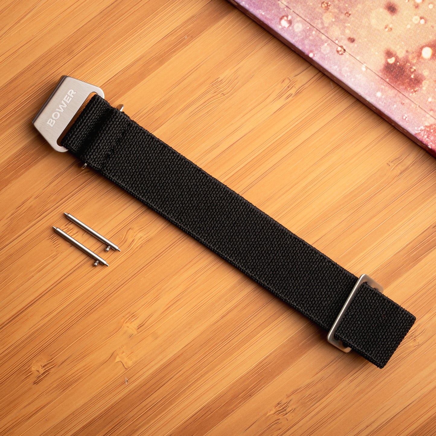 Black nylon watch strap by Bower with stainless steel hardware and quick-release spring bars, laid flat on a bamboo surface beside a speckled notebook. Durable, ocean-ready strap for outdoor wear.