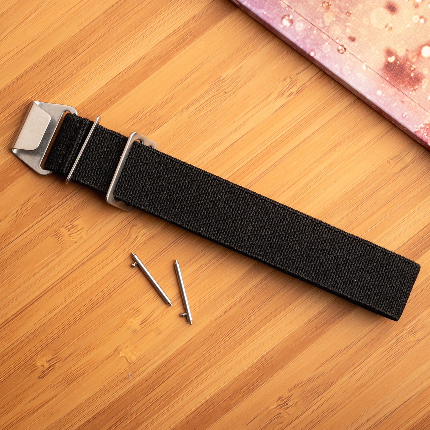 Black nylon watch strap by Bower with stainless steel hardware and quick-release spring bars, laid flat on a bamboo surface beside a speckled notebook. Durable, ocean-ready strap for outdoor wear.