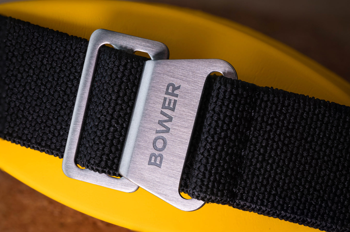 Close-up of Bower-branded stainless steel clasp on black nylon watch strap, resting on a yellow surface. Rugged and adjustable strap designed for ocean-ready timepieces.