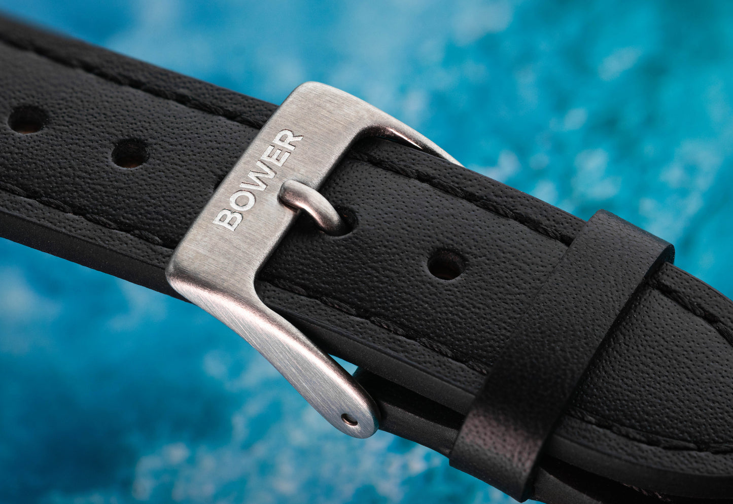 Close-up of Bower-branded stainless steel buckle on black leather watch strap, set against an ocean-inspired blue background. Elegant and durable strap designed for the Tide Seeker watch.