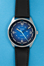 Studio image of a Bower tide watch with a deep blue textured dial, white hour markers, tide markings, and a black leather strap, displayed on a white background.