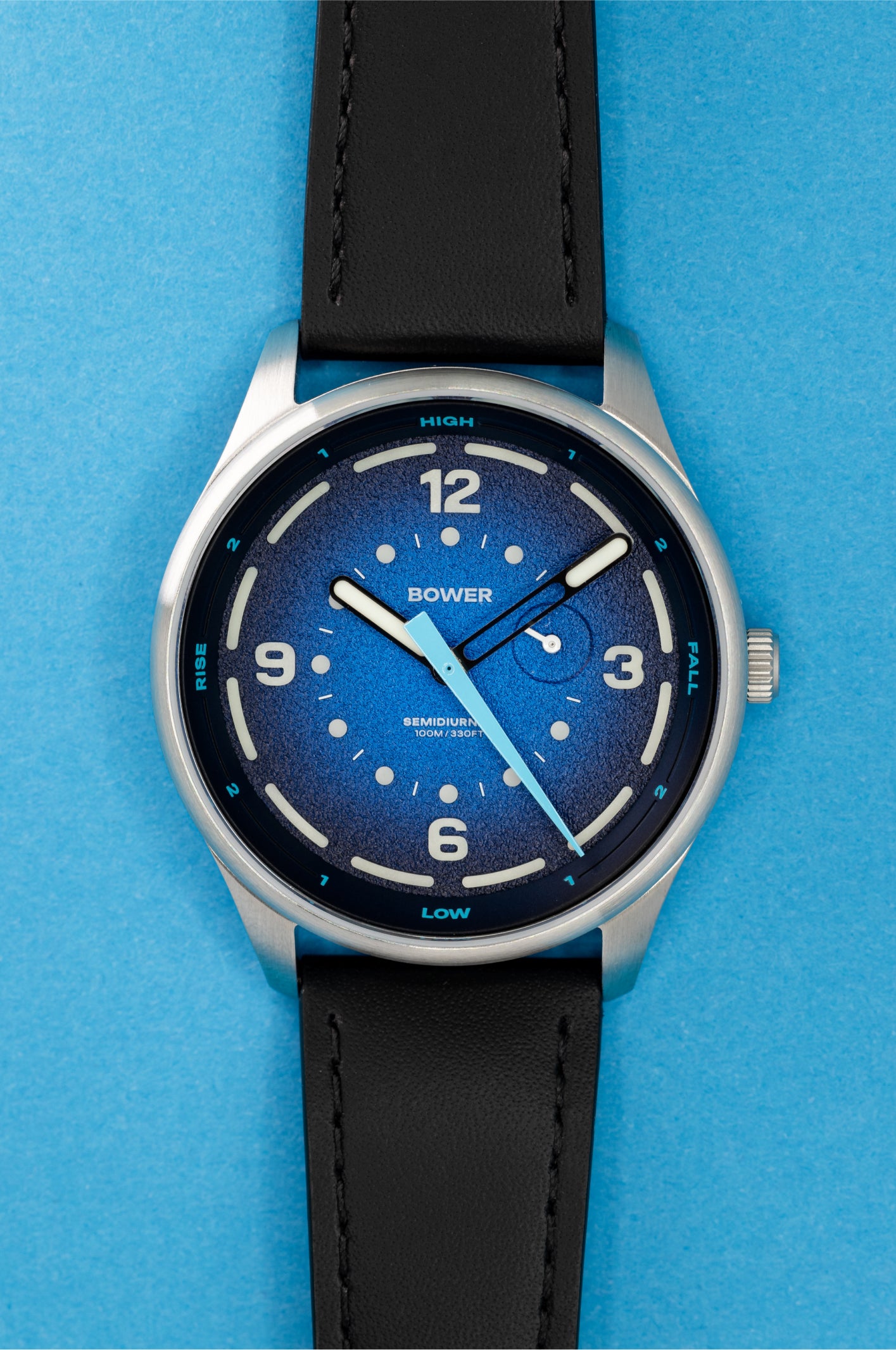Studio image of a Bower tide watch with a deep blue textured dial, white hour markers, tide markings, and a black leather strap, displayed on a white background.