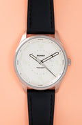 Studio image of a Bower tide watch with a coral white textured dial, white hour markers, tide markings, and a black leather strap, displayed on a white background.
