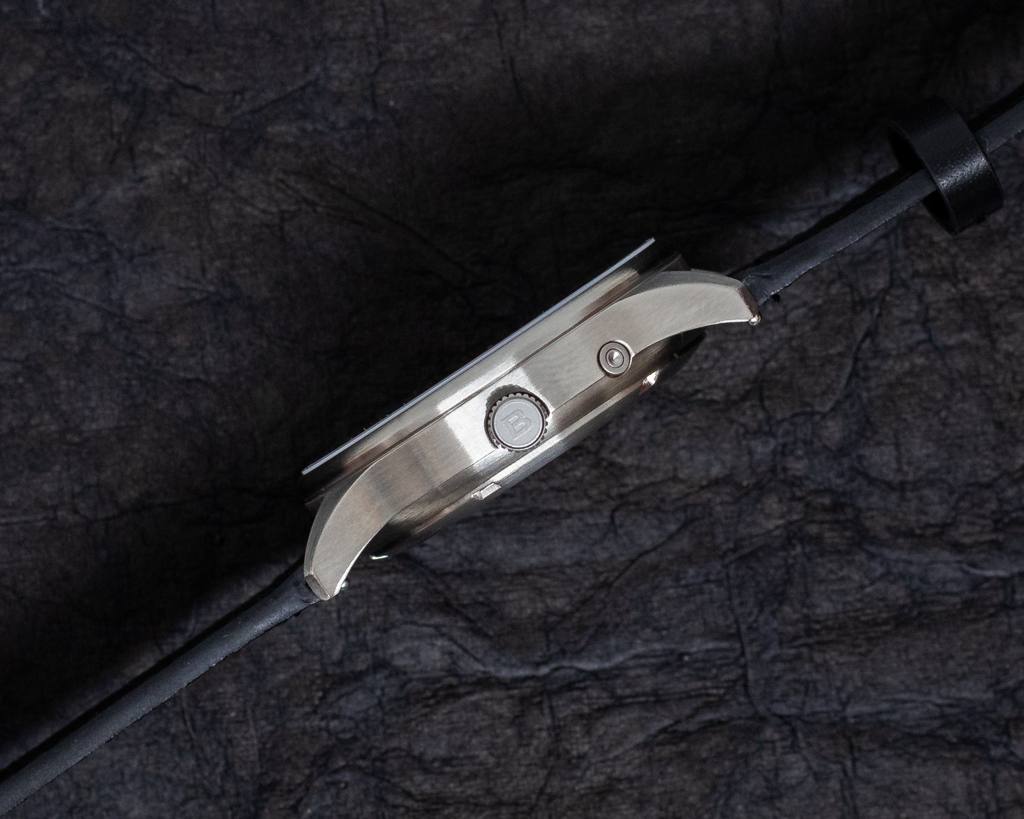 Side profile view of the Bower Tide Seeker watch showing the brushed stainless steel case, engraved crown, and recessed tide pusher. Set against a dark textured background, highlighting the watch’s slim and functional design.