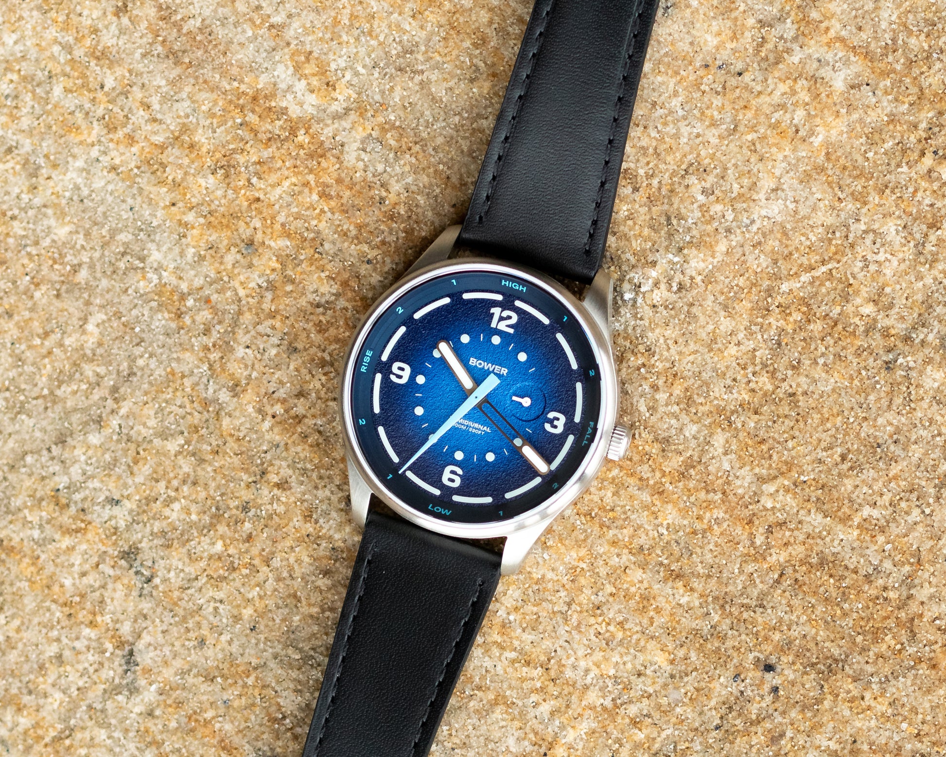 a blue dial bower watch laying flat on sand-like rocks by the ocean