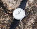 Close-up of a white Bower tide watch with a textured dial and black leather strap, resting on a weathered rock surface.