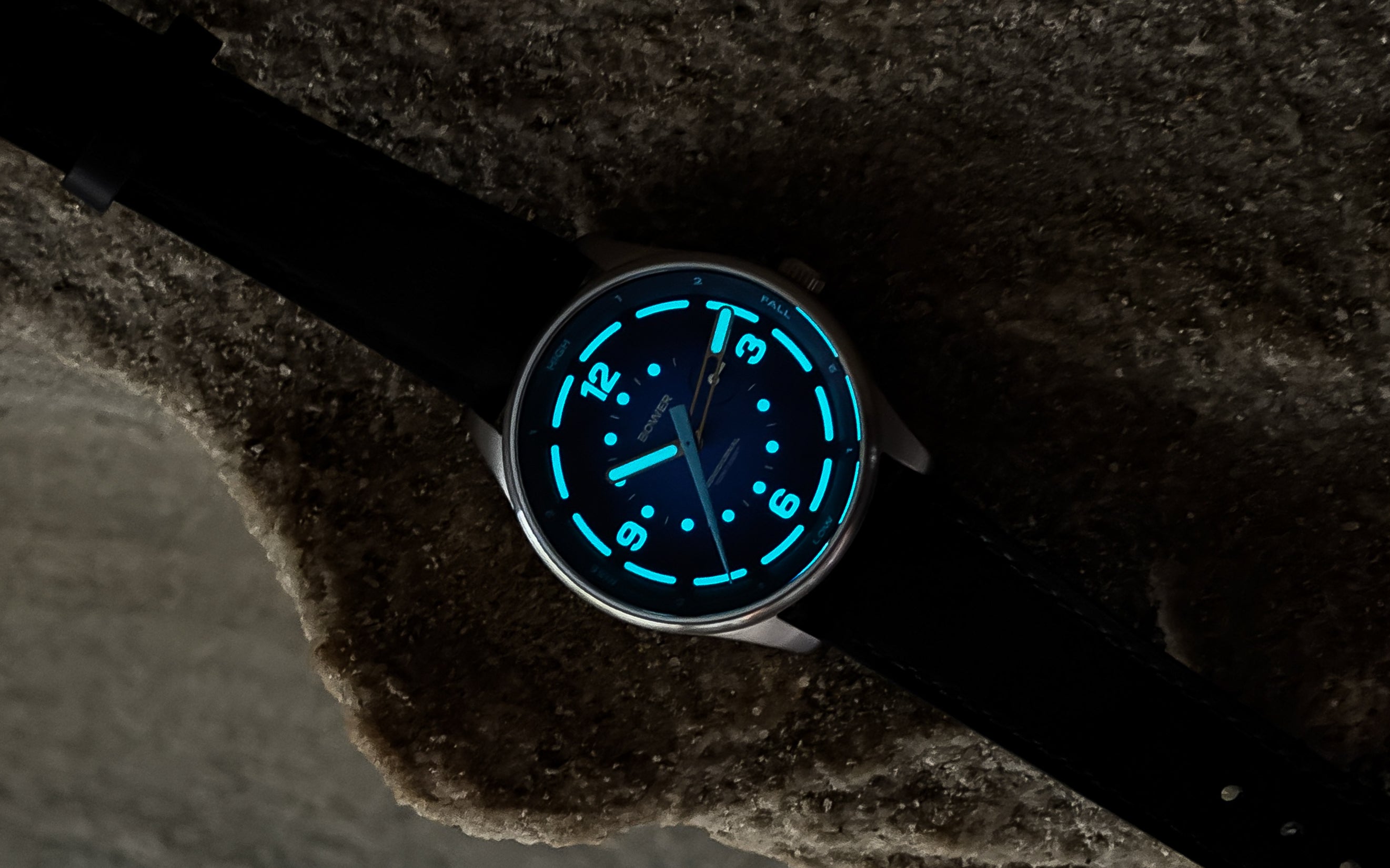 Bower Tide Seeker watch in low light, showing glowing blue luminescent markers using Swiss Super-LumiNova and hands on a Deep Blue dial. Resting on textured rock, the watch highlights its readability in dark environments and rugged outdoor styling.