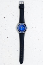 Flat lay of a Bower watch with a textured blue dial and black leather strap, displayed vertically on a speckled light background.
