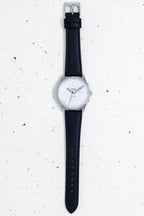 Flat lay of a Bower watch with a textured white dial and black leather strap, displayed vertically on a speckled light background.