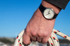 Close-up of a person’s hand gripping marine rope, wearing a Bower watch with a white dial and black strap, set against a clear blue sky and blurred waterfront background.