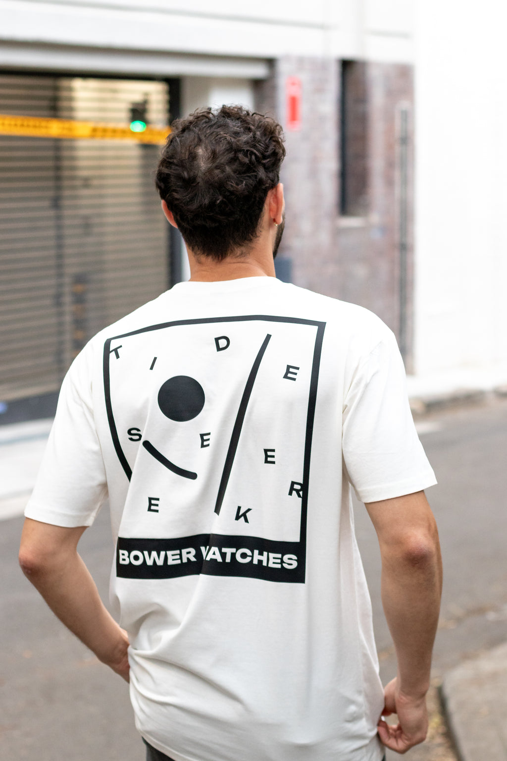 Person wearing a white Bower Watches T-shirt featuring a bold black graphic on the back that reads “TIDE SEEKER” with geometric tide-inspired shapes and “BOWER WATCHES” at the bottom, walking in the street in a fashion trendy loft district.