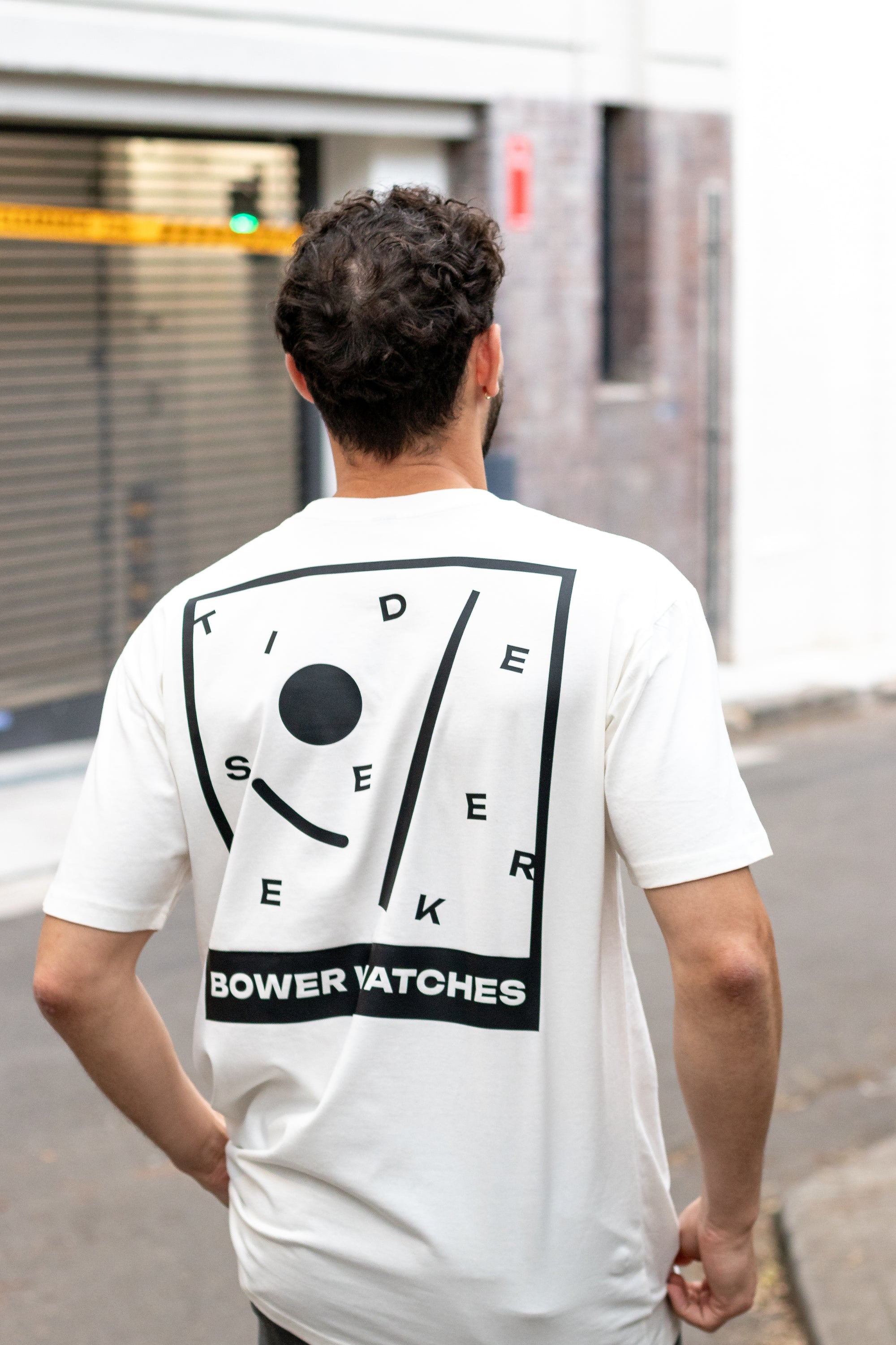 Person wearing a white Bower Watches T-shirt featuring a bold black graphic on the back that reads “TIDE SEEKER” with geometric tide-inspired shapes and “BOWER WATCHES” at the bottom, walking in the street in a fashion trendy loft district.