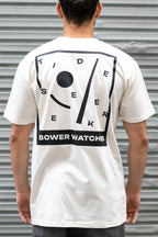 Person wearing a white Bower Watches T-shirt featuring a bold black graphic on the back that reads “TIDE SEEKER” with geometric tide-inspired shapes and “BOWER WATCHES” at the bottom, standing in front of a corrugated metal background.