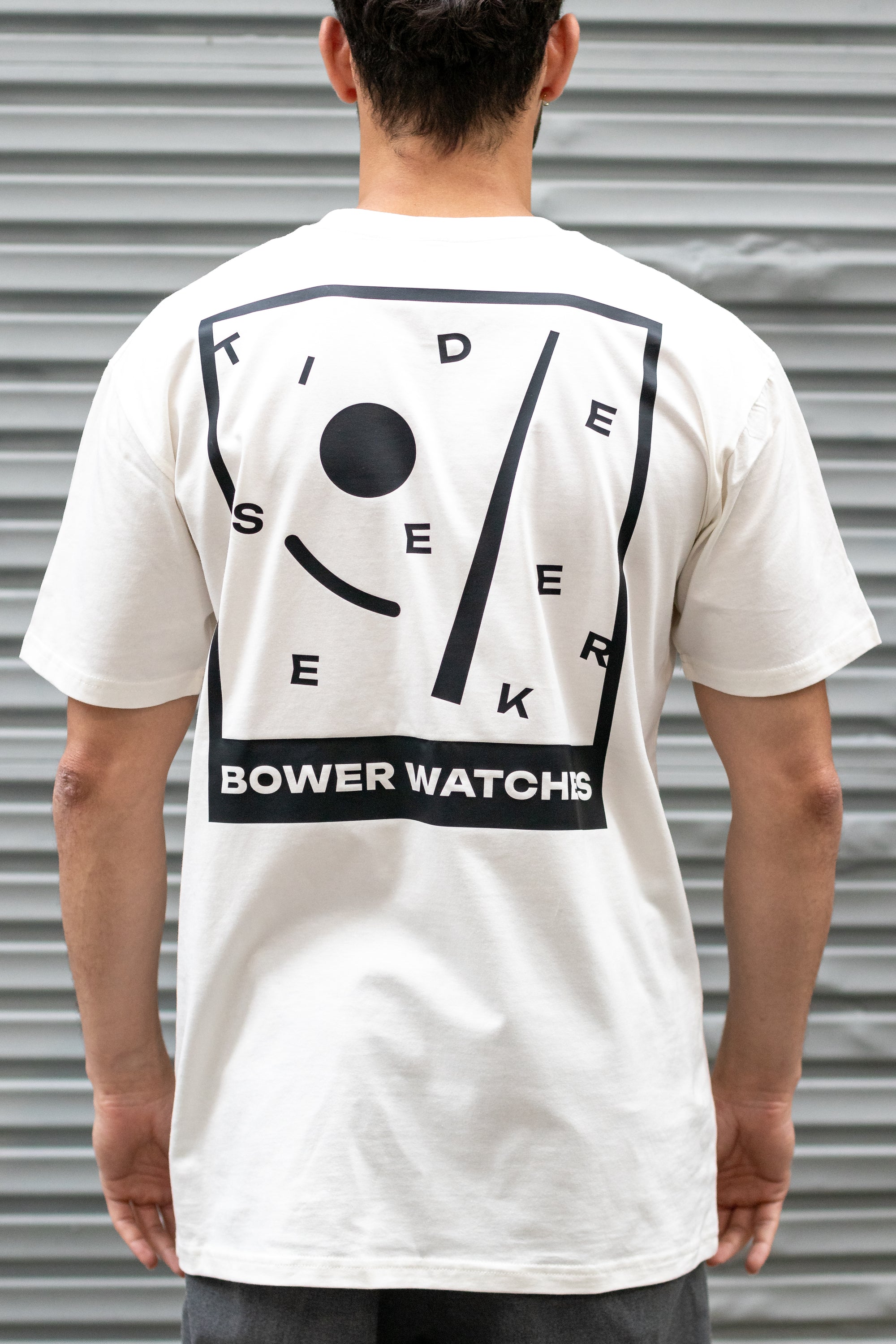 Person wearing a white Bower Watches T-shirt featuring a bold black graphic on the back that reads “TIDE SEEKER” with geometric tide-inspired shapes and “BOWER WATCHES” at the bottom, standing in front of a corrugated metal background.