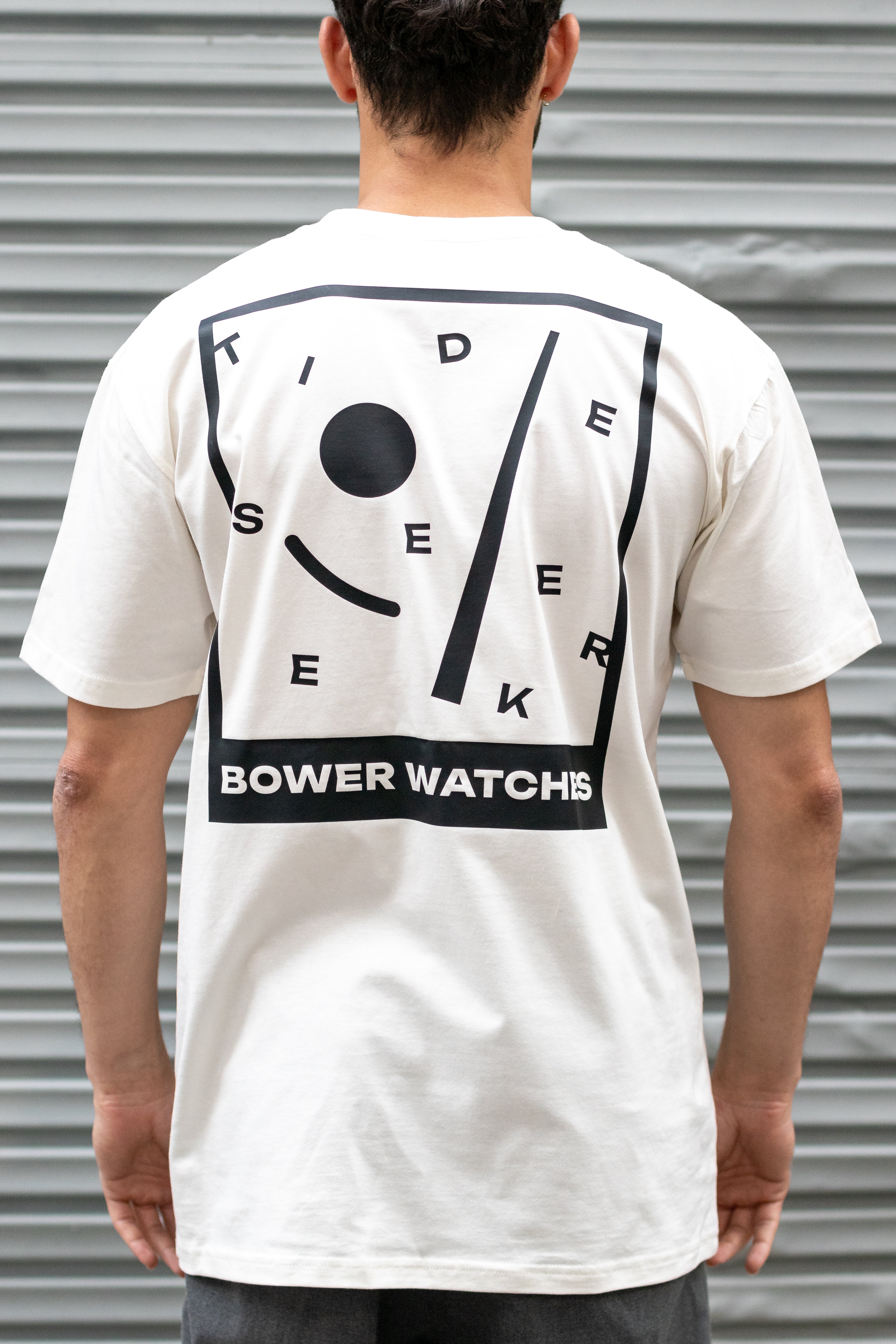 Person wearing a white Bower Watches T-shirt featuring a bold black graphic on the back that reads “TIDE SEEKER” with geometric tide-inspired shapes and “BOWER WATCHES” at the bottom, standing in front of a corrugated metal background.