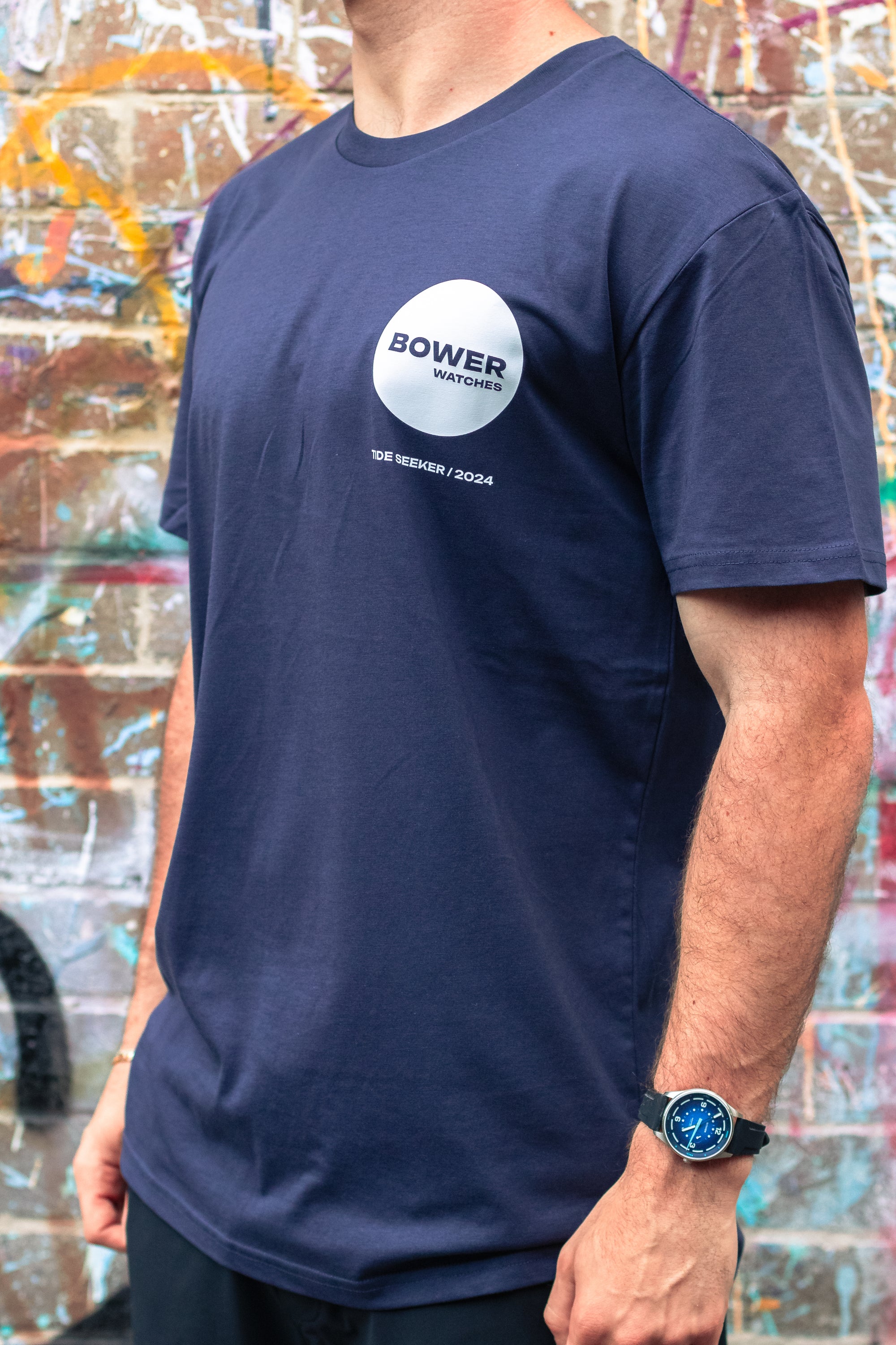 Man wearing a navy Bower Watches T-shirt with a bold white circle logo on the chest reading “BOWER WATCHES” and “TIDE SEEKER / 2024,” standing in front of a colorful graffiti-covered brick wall. He is also wearing a blue-dial Bower Tide Seeker watch on his wrist.