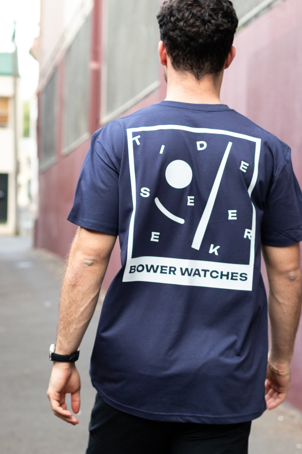Person wearing a navy blue T-shirt with a bold white graphic on the back that reads “TIDE SEEKER BOWER WATCHES.” The design features abstract tide symbols in a tilted frame. The person is walking down an urban alley, viewed from behind.