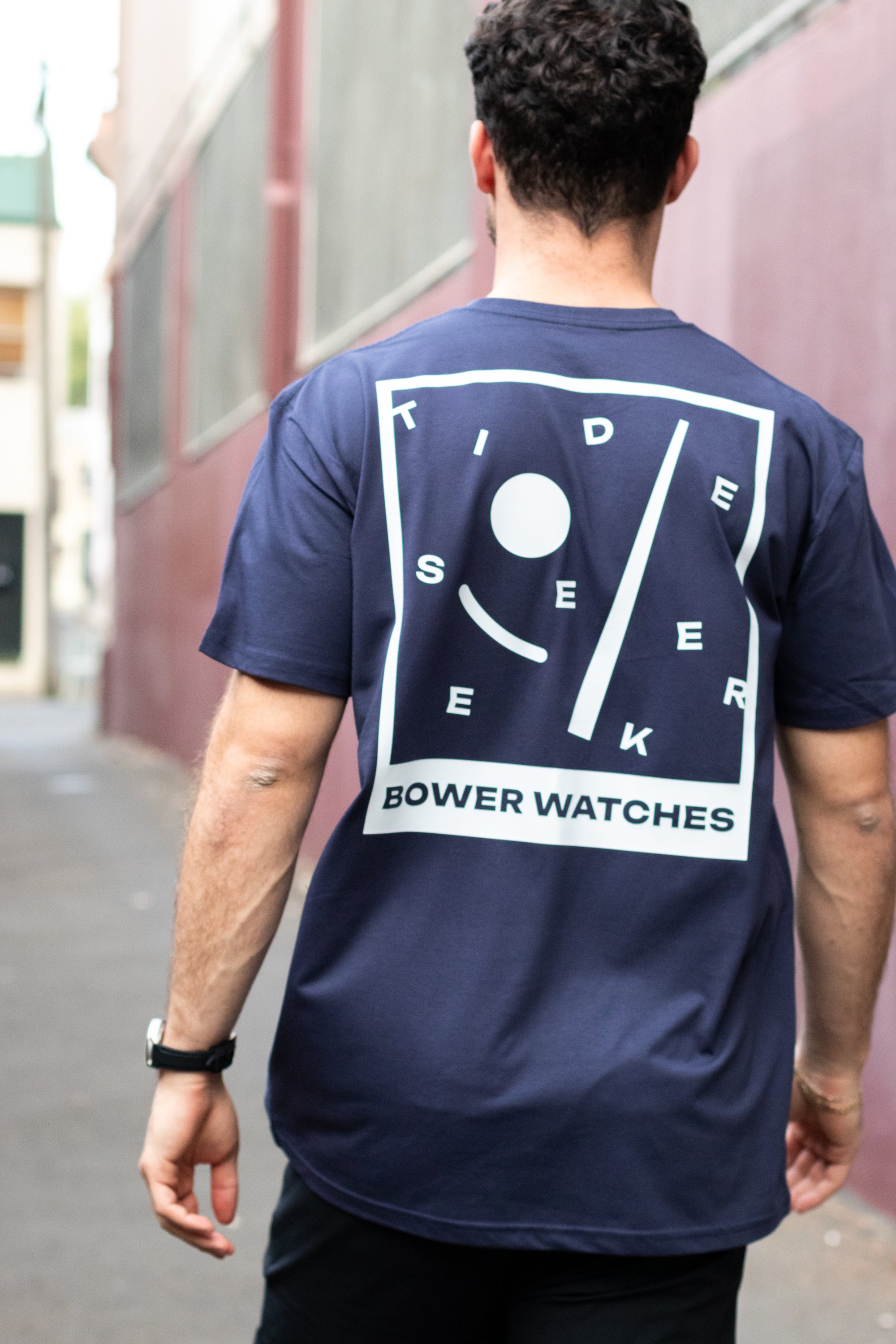 Person wearing a navy blue T-shirt with a bold white graphic on the back that reads “TIDE SEEKER BOWER WATCHES.” The design features abstract tide symbols in a tilted frame. The person is walking down an urban alley, viewed from behind.