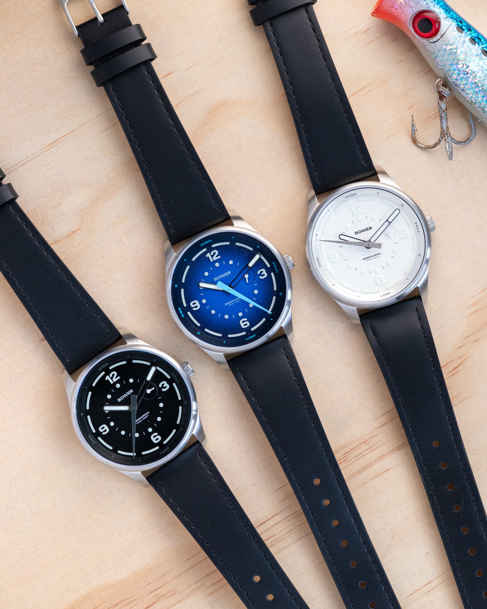 Three Bower Semidiurnal watches with black leather straps in black, blue, and white dials, displayed on a light wood surface next to a vintage fishing lure.