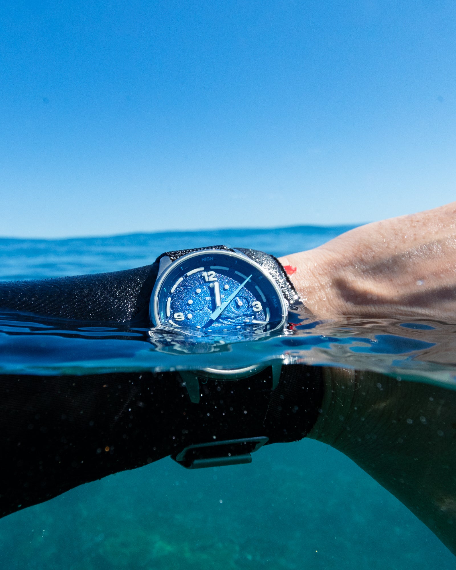 Close-up of a Bower blue tide watch partially submerged in ocean water, worn on wrist with a wetsuit under a clear blue sky.
