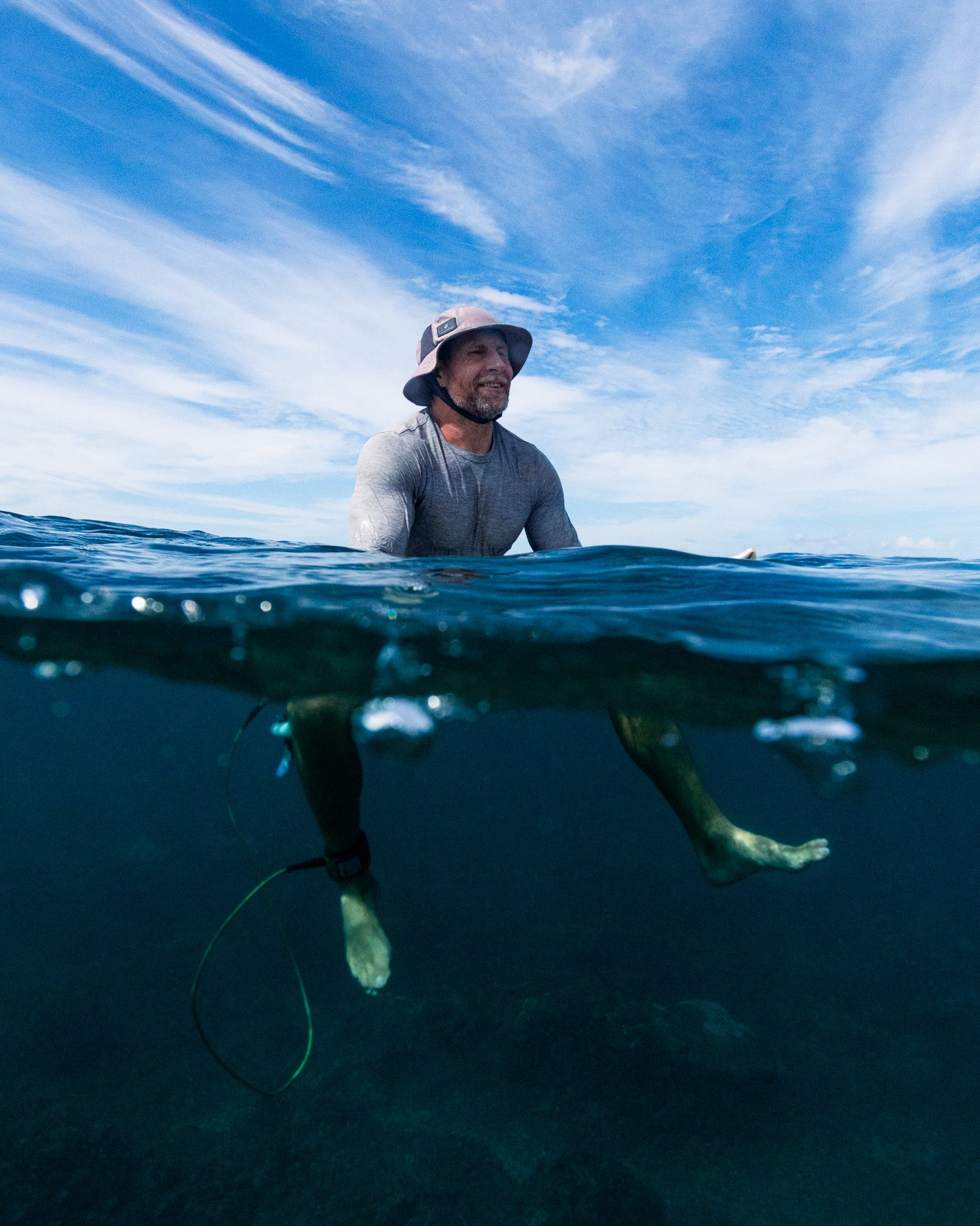 Surfer sitting in the ocean wearing a wide-brim hat and rash guard, captured during a Bower Watches field test in Fiji, with a clear split view above and below the waterline.