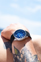 Close-up of a tattooed wrist wearing the Deep Blue Tide Seeker watch by Bower with a black fabric strap, against a bright, sunlit sky. Coastal lifestyle shot highlighting the watch’s tide-tracking design and textured dial.