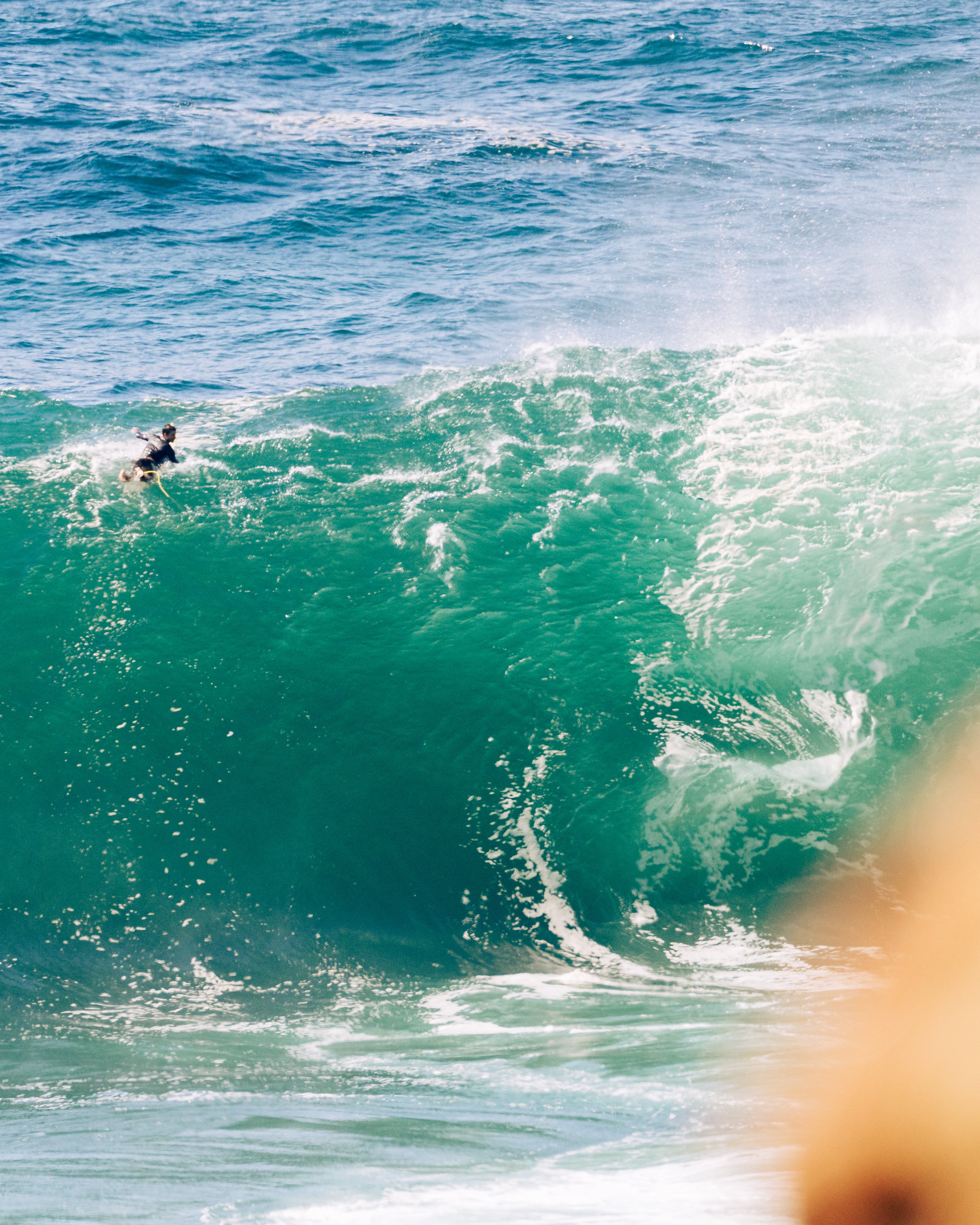 Surfer paddling into a massive green wave in open ocean, with spray breaking at the crest. Action shot capturing the scale and power of coastal surf conditions.
