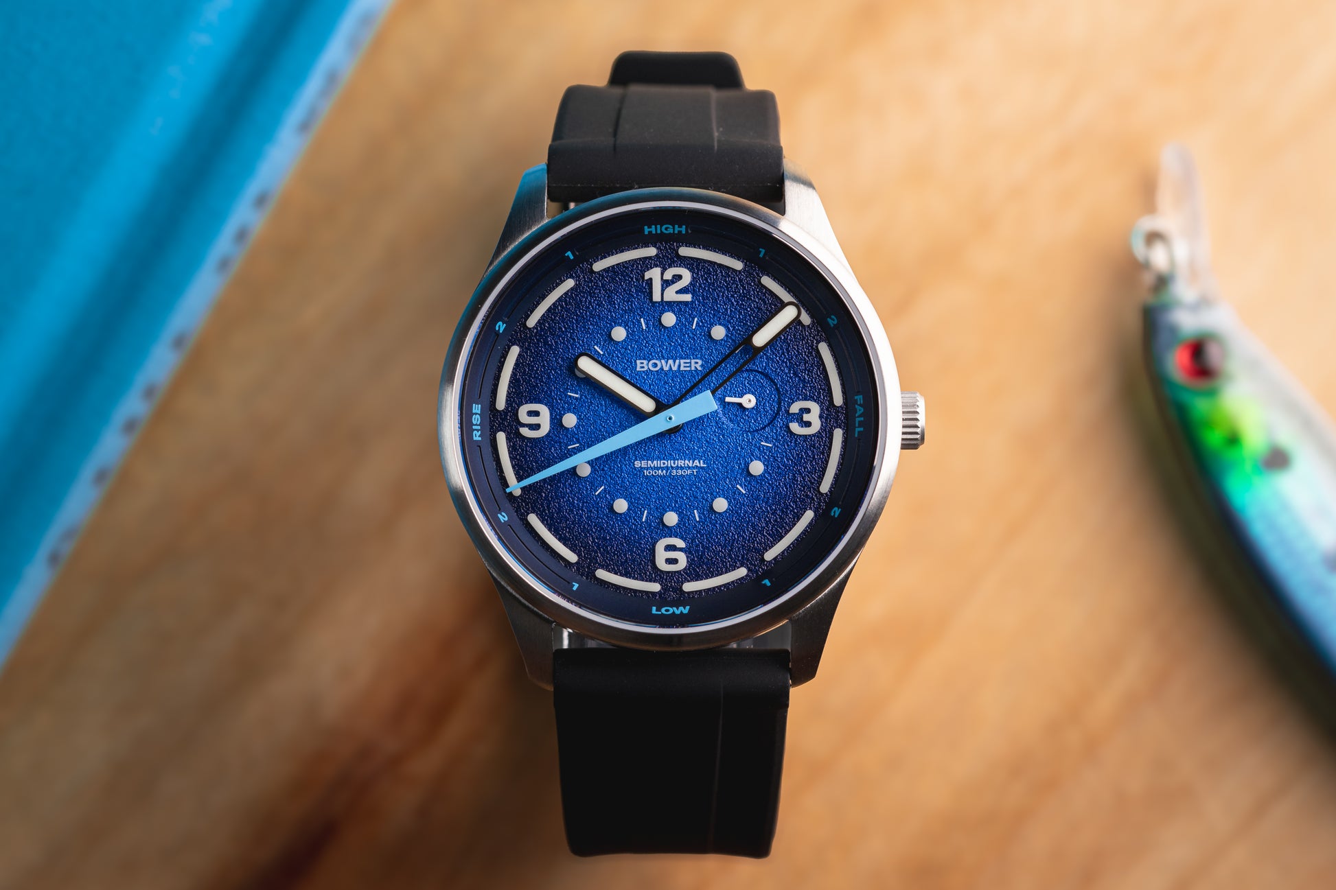 Deep Blue Tide Seeker watch by Bower with black rubber strap, displayed on wooden surface beside fishing lure and blue hardcover. Australian-designed tide watch for coastal and ocean adventures.