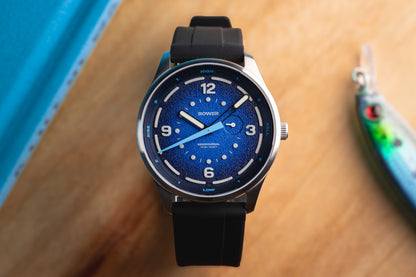 Deep Blue Tide Seeker watch by Bower with black rubber strap, displayed on wooden surface beside fishing lure and blue hardcover. Australian-designed tide watch for coastal and ocean adventures.
