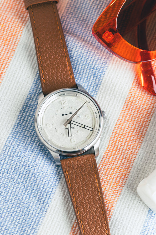 Coral White Tide Seeker watch by Bower with tan leather strap, styled on striped beach towel beside retro orange sunglasses and sunscreen. Designed in Australia for ocean lovers.
