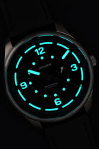 Close-up of the Bower semidiurnal tide watch glowing in the dark with bright aqua-blue lume, showing clear visibility of the hour, minute, second, and tide subdial hands.