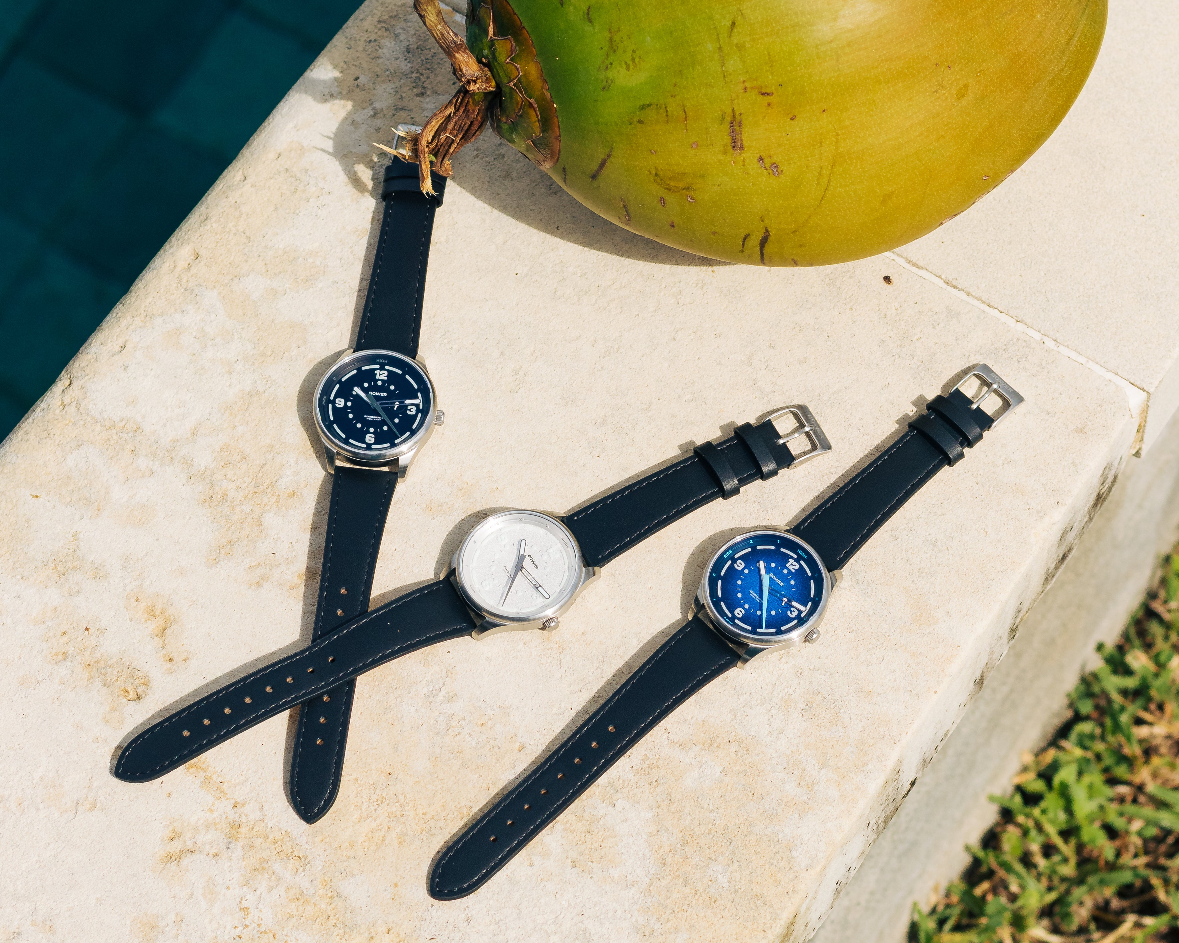 Three Bower Semidiurnal tide watches with black, white, and blue dials arranged on a stone ledge next to a green coconut and a swimming pool.