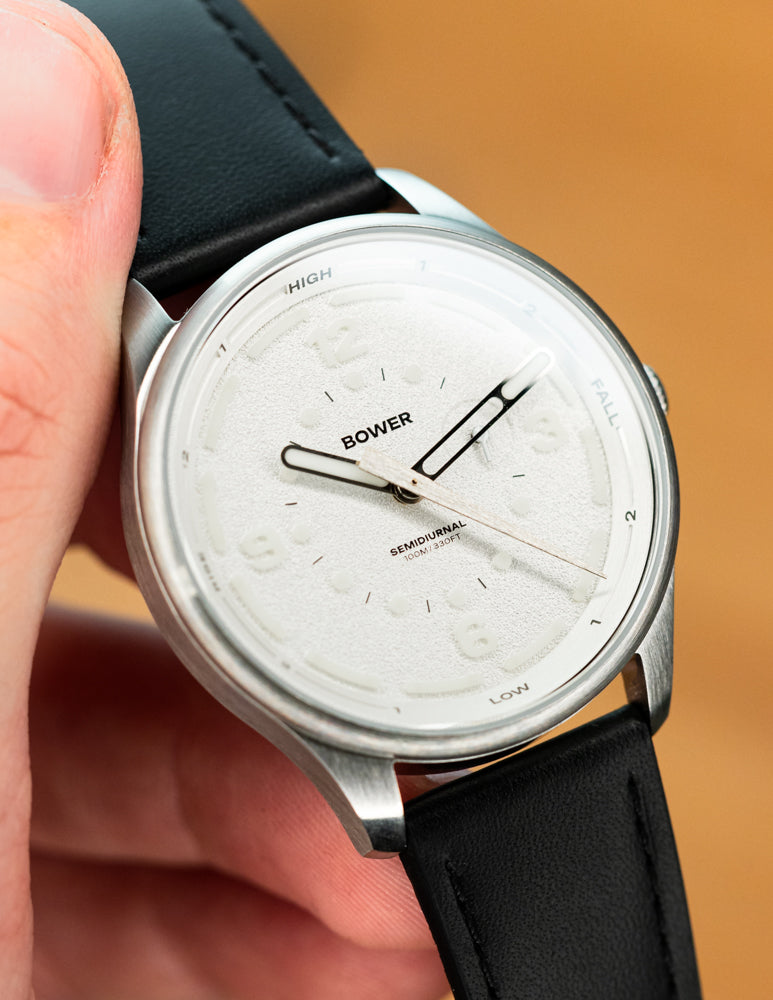 Close-up of the Bower Tide Seeker watch in white, showing the semidiurnal tide hand, embossed markers, and brushed stainless steel case held in hand.