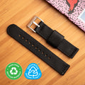 Black recycled watch strap by Bower with brushed stainless steel buckle, laid on a bamboo surface beside a colourful notebook. Labeled as made from recycled materials and prevented ocean plastic.
