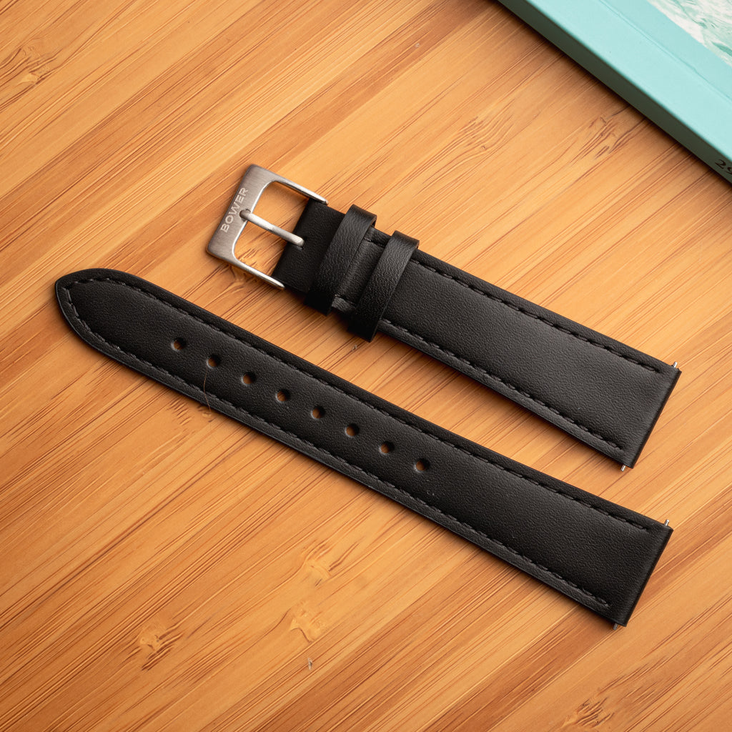 Black leather watch strap by Bower with brushed stainless steel buckle, displayed on a bamboo surface next to a turquoise notebook. Refined and versatile strap designed for everyday or dress wear.