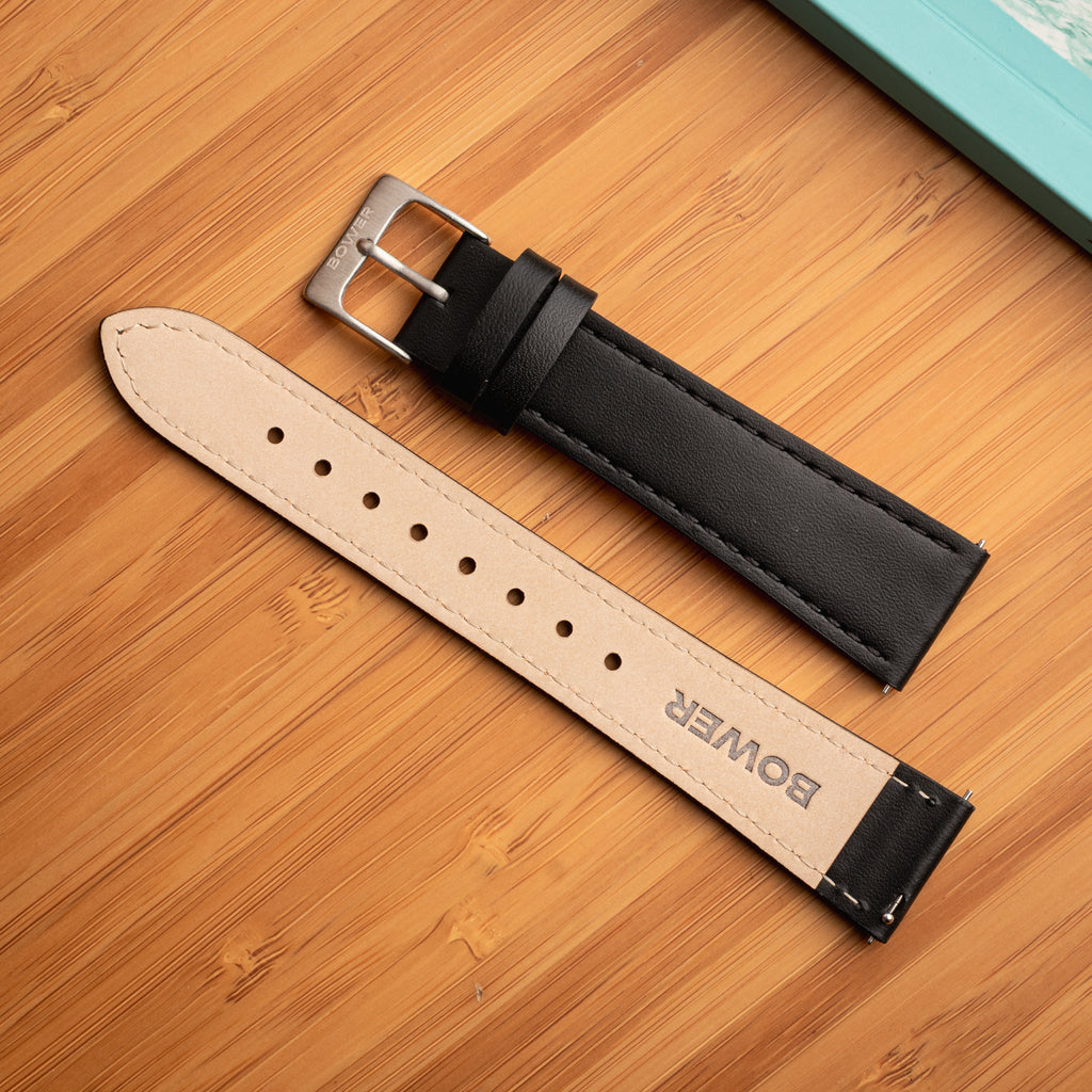 Underside of black leather watch strap by Bower, showing beige lining with BOWER logo and stainless steel buckle. Displayed on a bamboo surface beside a turquoise notebook.