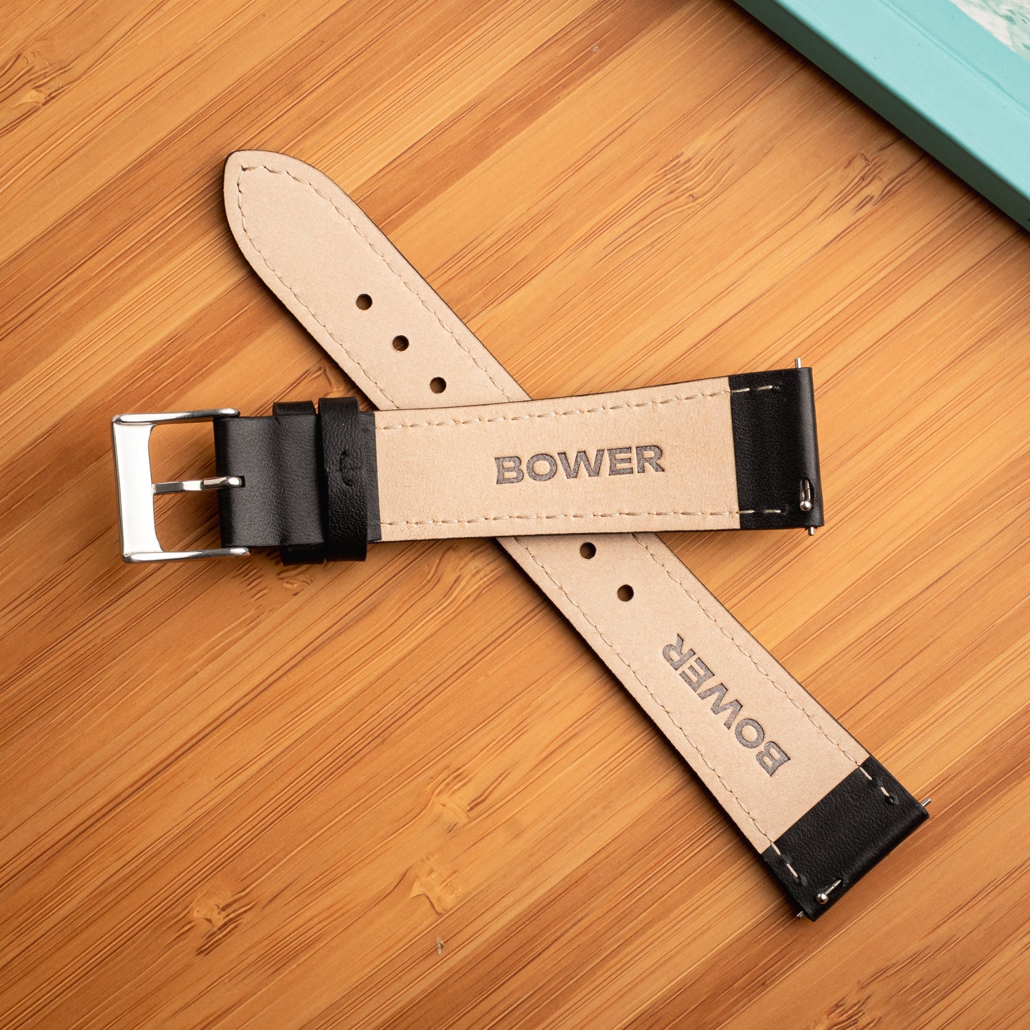 Underside of black leather watch strap by Bower, showing beige lining with BOWER logo and stainless steel buckle. Displayed on a bamboo surface beside a turquoise notebook.