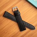 Black leather watch strap by Bower with brushed stainless steel buckle, displayed on a bamboo surface next to a turquoise notebook. Refined and versatile strap designed for everyday or dress wear.