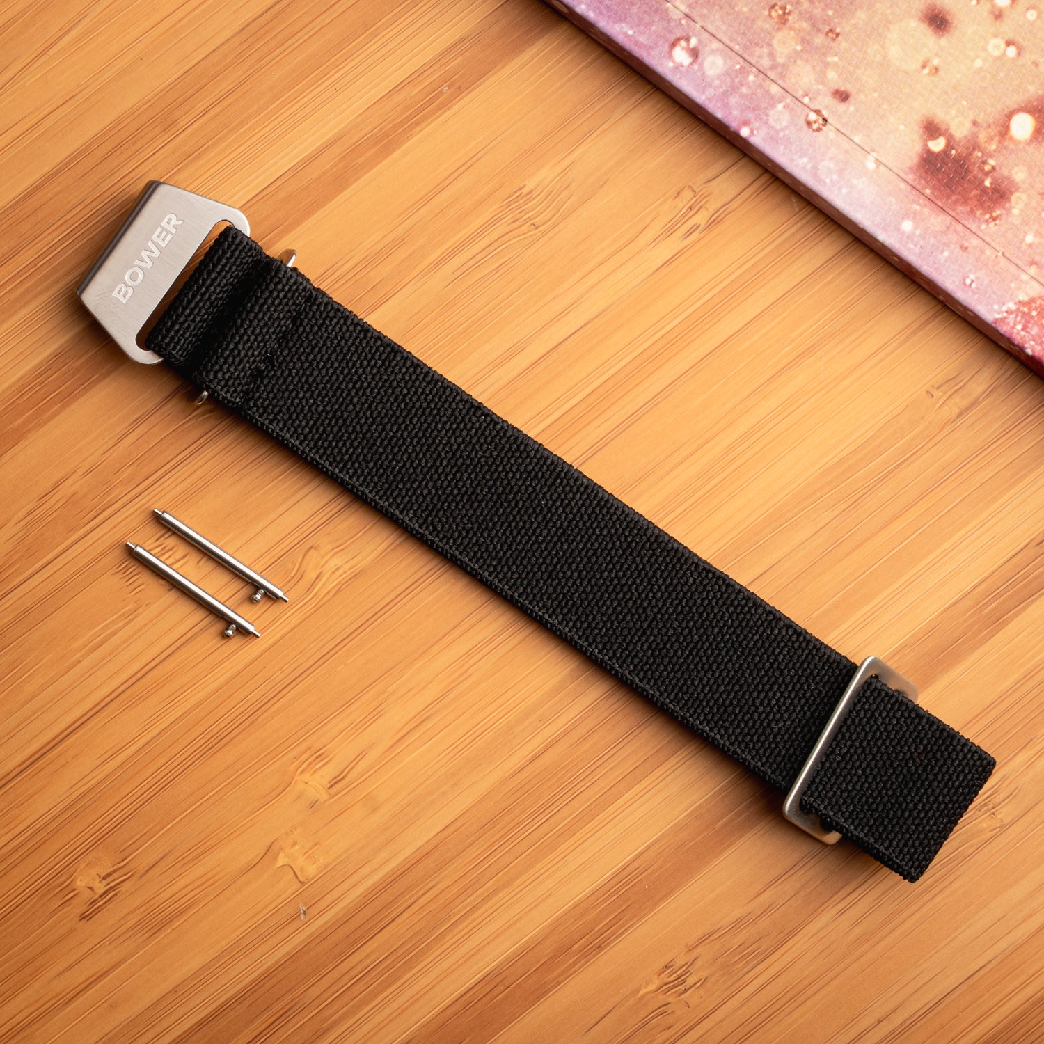 Black nylon watch strap by Bower with stainless steel hardware and quick-release spring bars, laid flat on a bamboo surface beside a speckled notebook. Durable, ocean-ready strap for outdoor wear.