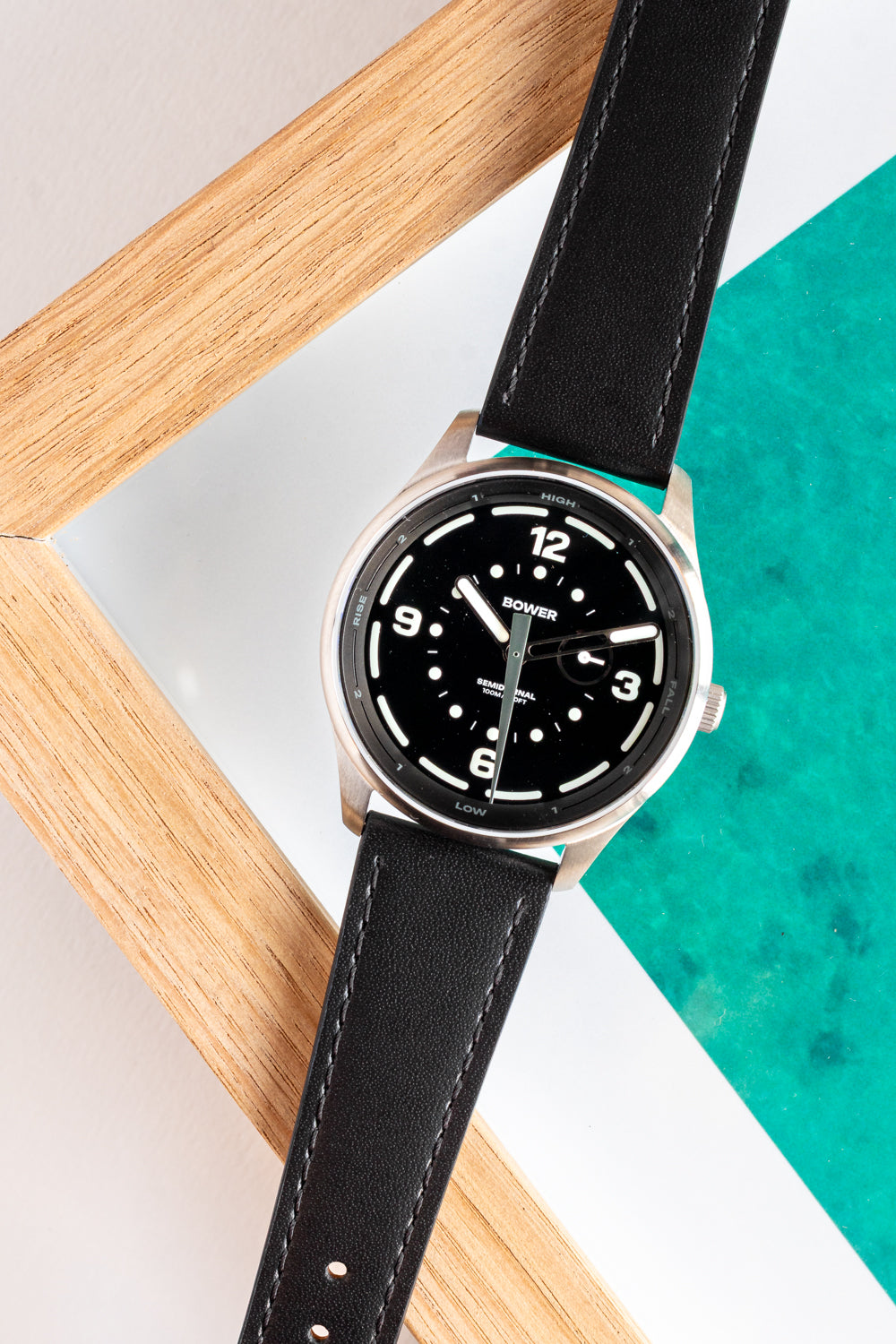 Midnight Black Tide Seeker watch by Bower with black leather strap, displayed diagonally on a wooden frame and teal ocean-inspired background. Minimalist tide-tracking timepiece designed for coastal living.