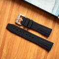 Black silicone watch strap by Bower with brushed stainless steel buckle, displayed on a bamboo surface next to a light blue notebook. Durable and water-resistant strap ideal for active coastal use.