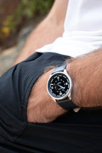 Man wearing a Midnight Black Tide Seeker watch by Bower with black leather strap, resting hand in pocket while seated outdoors. Casual lifestyle shot showcasing the watch in everyday use.