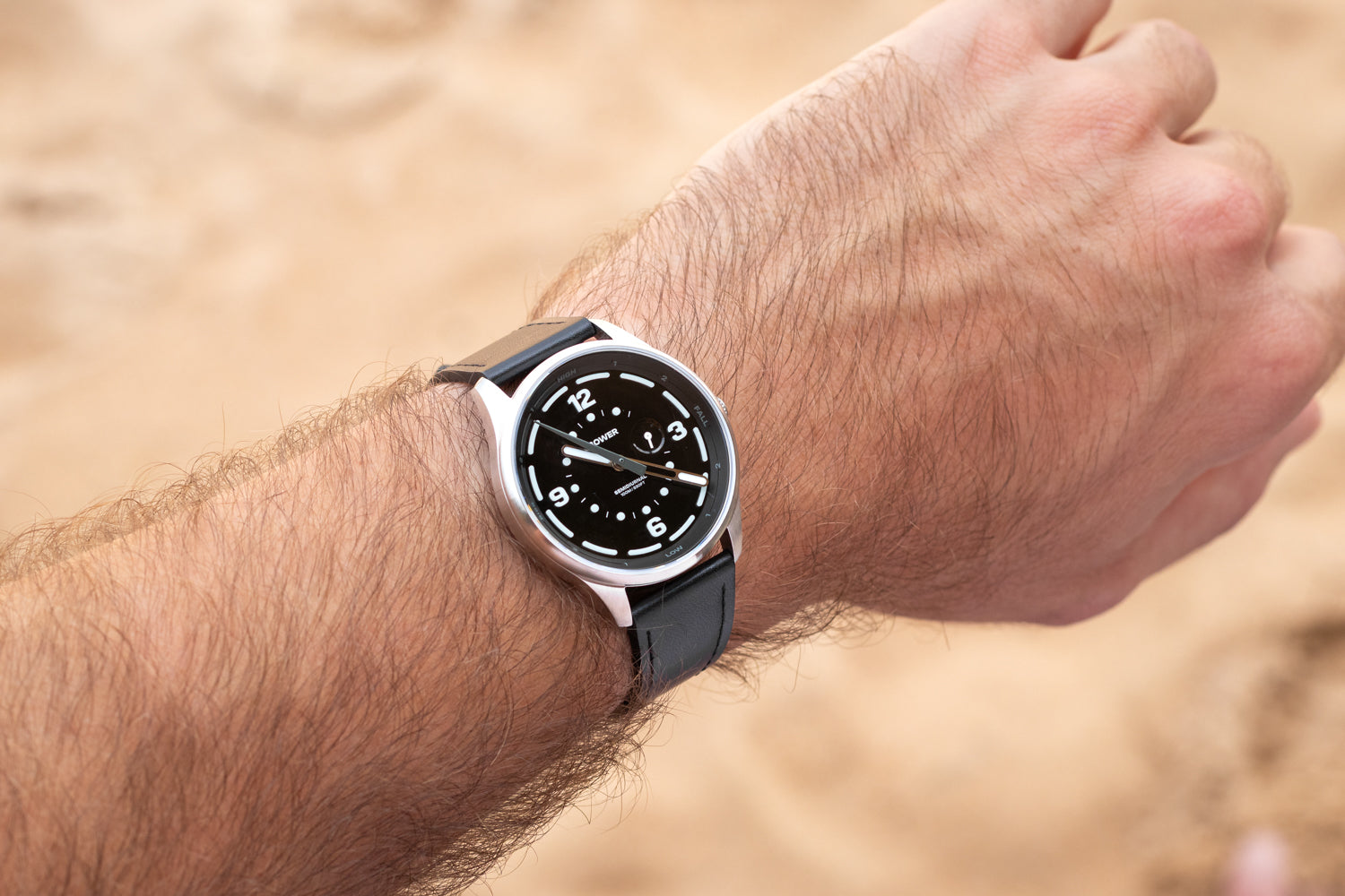 Midnight Black Tide Seeker watch by Bower worn on wrist, featuring black leather strap and luminous hands, with a sandy beach background. Coastal lifestyle timepiece for ocean explorers.