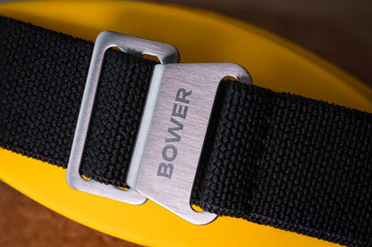 Close-up of Bower-branded stainless steel clasp on black nylon watch strap, resting on a yellow surface. Rugged and adjustable strap designed for ocean-ready timepieces.