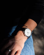 Close-up of a person’s wrist wearing a Bower watch with a white textured dial and black strap, paired with a minimalist silver cuff bracelet, captured in natural light.