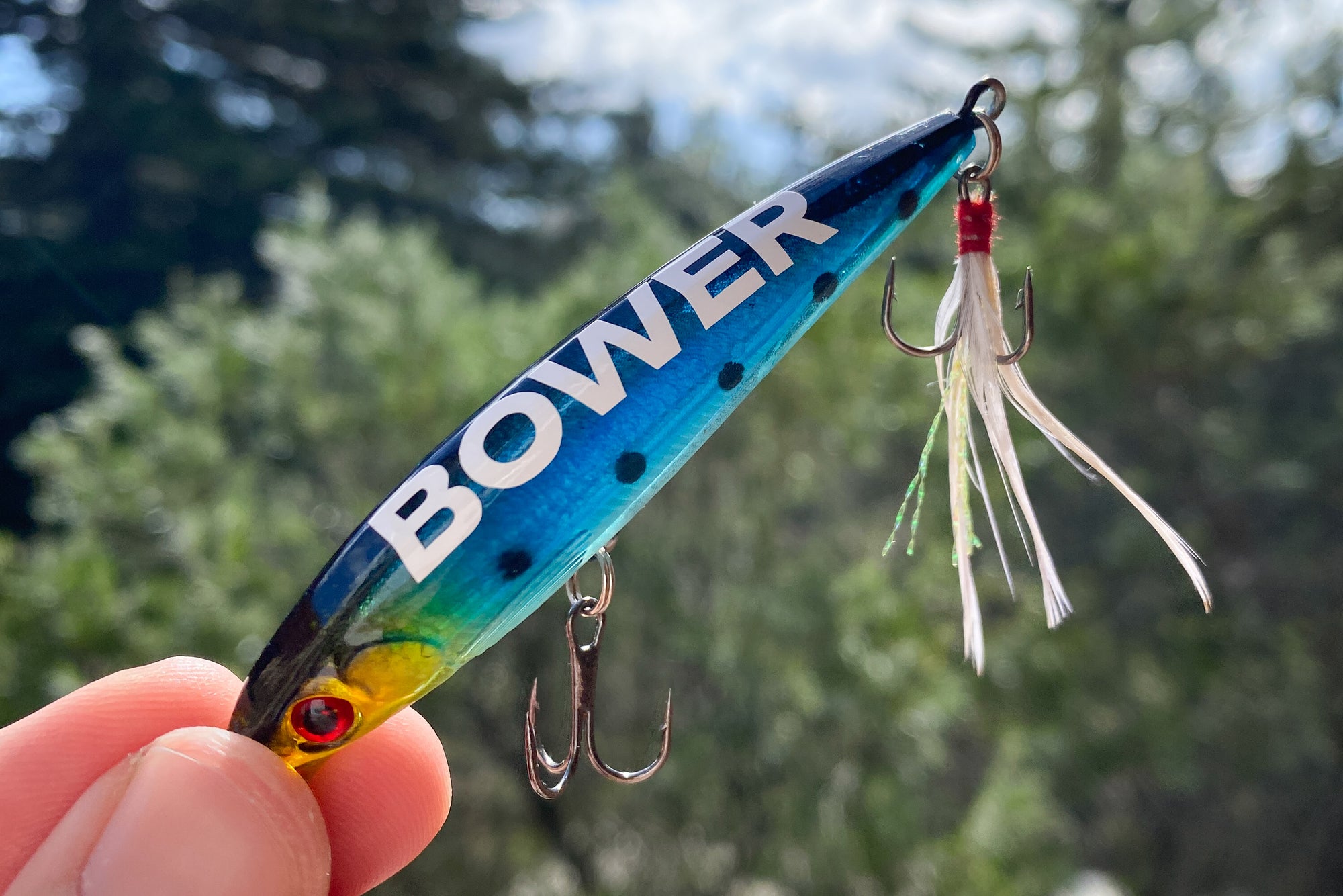 Hand holding a colorful fishing lure with the word “BOWER” printed in bold white letters. The lure has a gradient finish with blue, green, and yellow tones, black spots, and twin treble hooks. Trees and sky are softly blurred in the background.